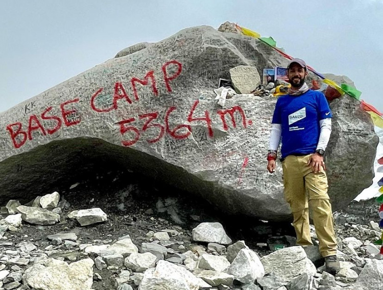 Karan Saberwal stands in his Baird shirt next to a large rock with "Base Camp 5364m." spray painted on it