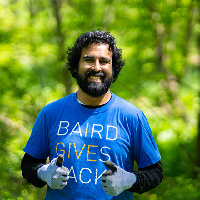 Baird associate standing outdoors wearing a Baird Gives Back shirt and giving a double thumbs up.