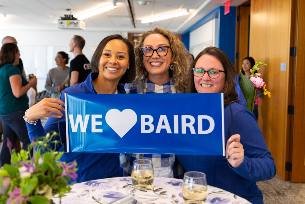 Three women holding a We Love Baird sign while attending the Baird Annual Meeting