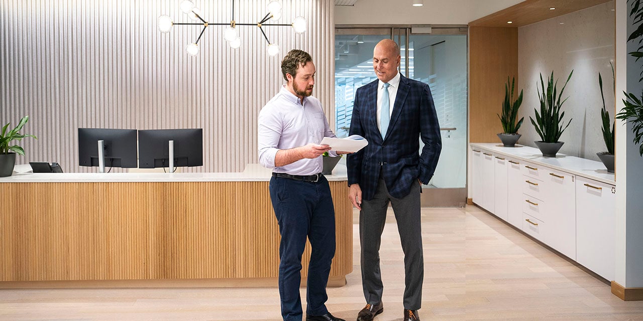 Two men standing in an office lobby reviewing paperwork that is being held by one of the individuals.