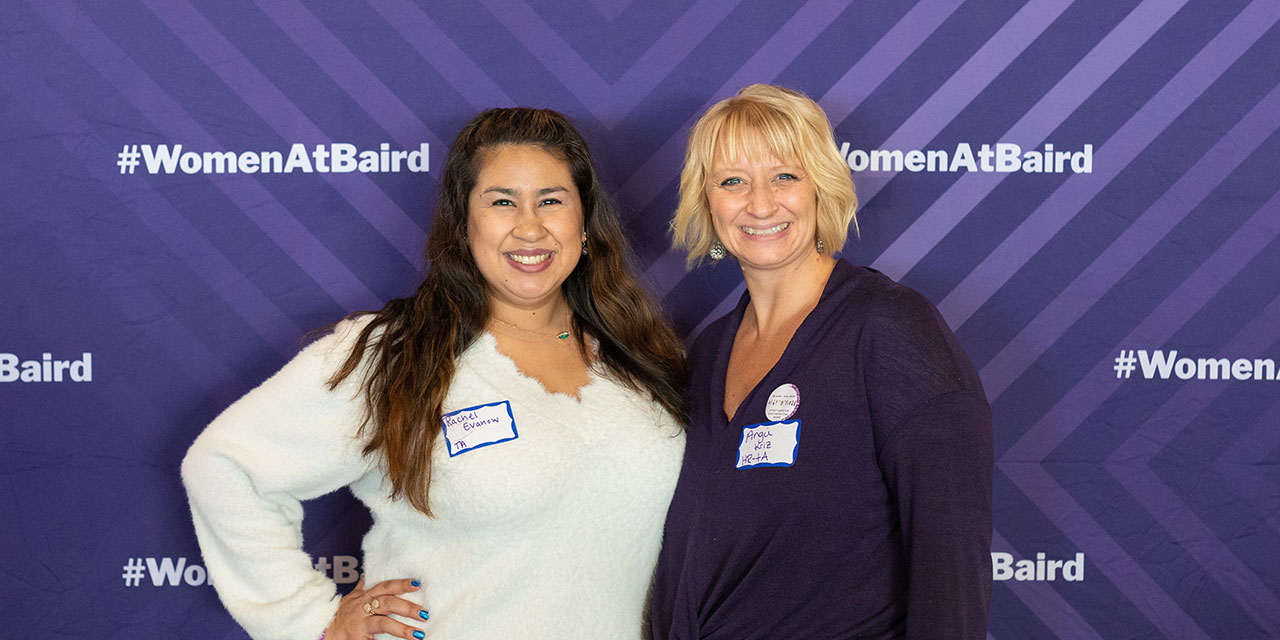 Two Baird associates in front of a purple banner with #womenatbaird hashtags