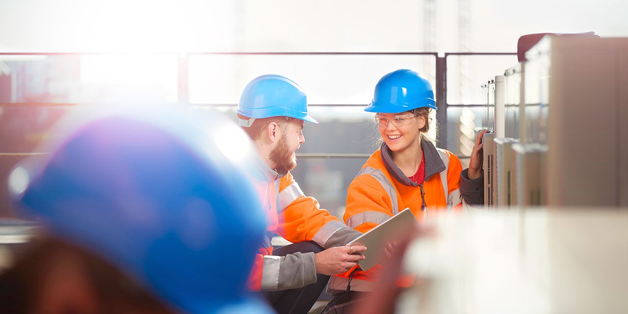 Workers in an industrial setting wearing safety vests and blue hardhats. 